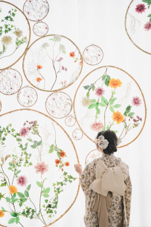 STUDIO ARCららぽーと湘南平塚店で撮影された七五三の記念写真。花柄の着物に生成り色の大きなリボン結びの帯を合わせ、白い花の髪飾りをつけた女の子が後ろ姿で、円形のフープに生けられた色とりどりの花や刺繍のオブジェを見上げている