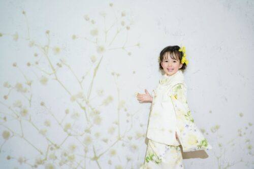 STUDIO ARCイオン相模原店で撮影された、着物姿の女の子の記念写真。黄色い花の髪飾りをつけ、白地に緑や黄色の花柄をあしらった被布姿で、片手を軽く広げて笑顔を見せている。背景には草花の絵柄が描かれた淡い色合いの壁が広がっている