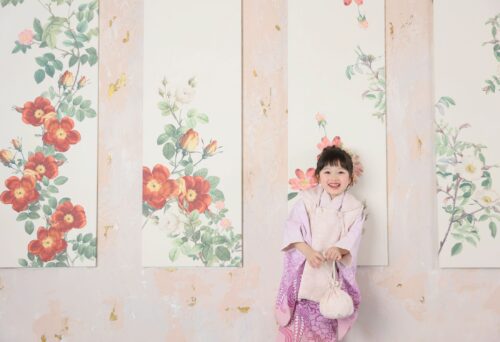 STUDIO ARCららぽーと湘南平塚店で撮影された七五三の記念写真。ピンクと紫のグラデーションの着物に白い被布を合わせ、巾着袋を手に持った女の子が笑顔で立っている。背景にはバラの花が描かれたパネルが並ぶ壁が広がっている