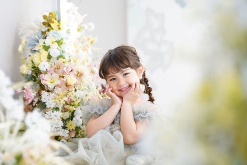 ららぽーと立川立飛店で撮影された女の子の記念写真。編み込みヘアに水色のチュールドレスを着て、両手を頬に添えて笑顔を見せている。隣にはピンクや黄色、白の花々が飾られている