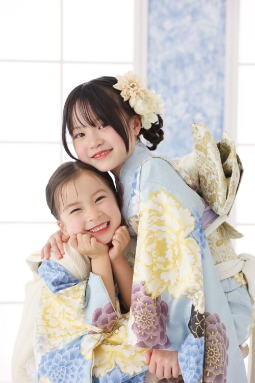STUDIO ARCららぽーと堺店で撮影された七五三の記念写真。水色に牡丹の花模様が描かれた振袖に白い花飾りをつけた姉が、水色の着物を着た妹を後ろから優しく抱きしめ、二人とも満面の笑顔を見せている