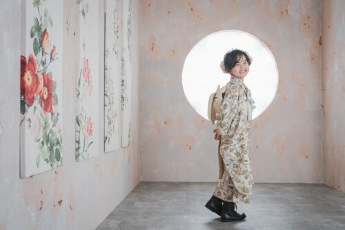 STUDIO ARCららぽーと湘南平塚店で撮影された七五三の記念写真。生成り色に花柄をあしらった着物に黒いブーツを合わせた女の子が、満月のような円形の光を背にして振り返り、満面の笑顔を見せている。左手にはバラの花が描かれたパネルが並ぶ壁が続いている