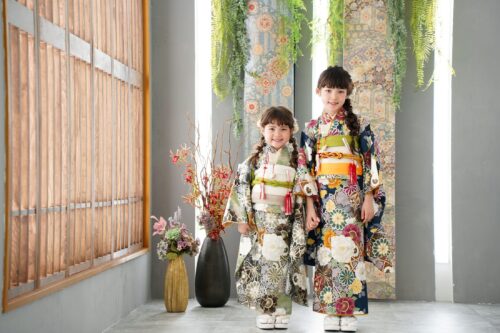 ららぽーと立川立飛店で撮影された、着物姿の姉妹の記念写真。緑色の帯を締めた小さな女の子と、紺色の花柄の振袖に橙色の帯を合わせた姉が手をつなぎ、笑顔でこちらを見ている。背景には格子状の木製パネルと、花柄のタペストリー、緑の植物が飾られている