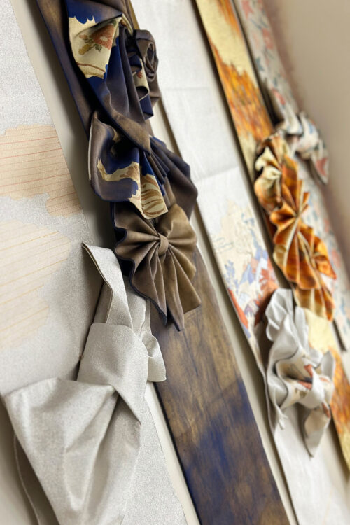 STUDIO ARC心斎橋店の店内ディスプレイ。着物の帯地で作られたお花のコサージュやリボン飾りが、金糸や紺、オレンジなど華やかな柄の帯とともに壁面に並べられている、和の伝統美を感じる展示コーナー。