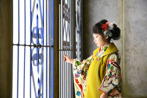七五三の記念撮影で、からし色の被布に花柄の着物を合わせた女の子が、アンティークな格子窓に手を添えて横顔で外を眺めている写真