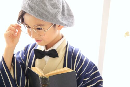 STUDIO ARC錦糸町PARCO店で撮影された、レトロな装いの男の子の記念写真。グレーのベレー帽と丸い眼鏡、紺と白のストライプ羽織に黒の蝶ネクタイを合わせ、古びた本を手に読書をするポーズで写っている