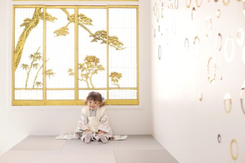 STUDIO ARCららぽーと門真店で撮影された七五三の記念写真。生成り色に花柄をあしらった着物に白い被布を合わせた女の子が、畳の上に足を崩して座り笑顔を見せている。背景には松や竹、牡丹が描かれた金色の襖と、輪っかの模様が飾られた白い壁が広がっている