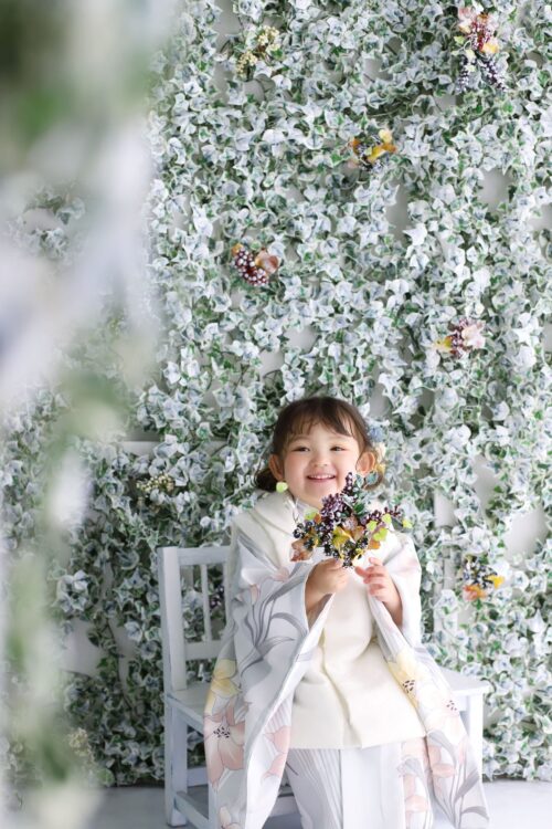 STUDIO ARCららぽーと門真店で撮影された七五三の記念写真。白地に花柄をあしらった着物を着た女の子が、白い椅子に座って木の実をあしらった小さなブーケを手に満面の笑顔を見せている。背景には斑入りの緑の葉が一面に垂れ下がる装飾壁が広がっている