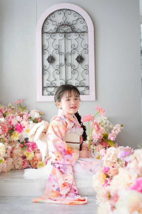 STUDIO ARC錦糸町PARCO店で撮影された、着物姿の女の子の記念写真。ピンクや白の花柄振袖に生成り色の帯を合わせ、ピンクのガーベラを手に微笑んでいる。背景にはアイアン装飾のアーチ型パネルと、ピンクや白の花々があふれるフラワーコーナーが飾られている