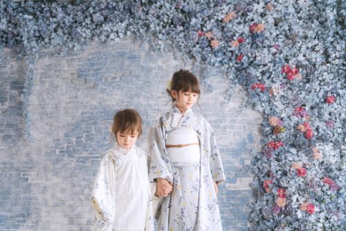 STUDIO ARC横浜店で撮影された、着物姿の姉妹の記念写真。白地に小花模様の着物を着た二人が手をつなぎ、青いレンガ調の壁と、あじさいや小花で彩られたブルーとピンクの花壁を背景に立っている