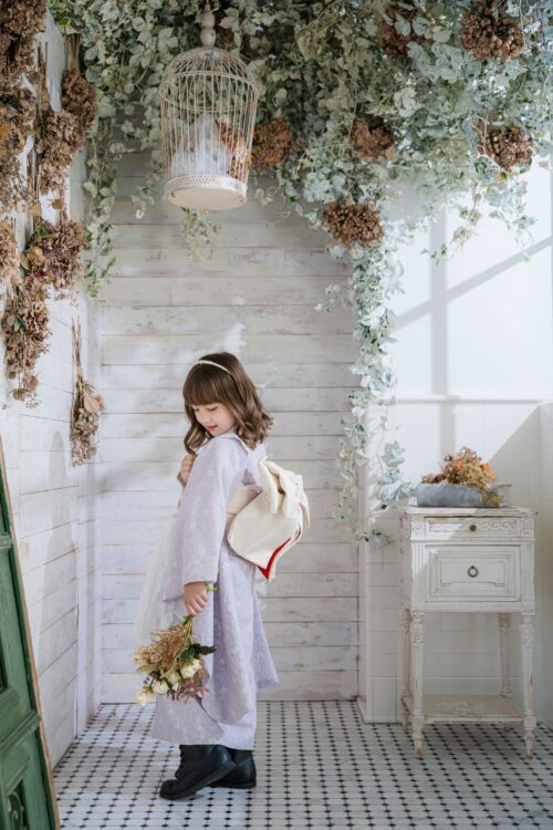 STUDIO ARCセンター北店で撮影された、着物姿の女の子の記念写真。淡いラベンダー色の着物に黒いブーツを合わせ、ドライフラワーのブーケを手に振り返る姿勢で立っている。頭上には鳥かご風のランタンとグリーンが吊るされ、白いアンティーク調の棚が飾られている
