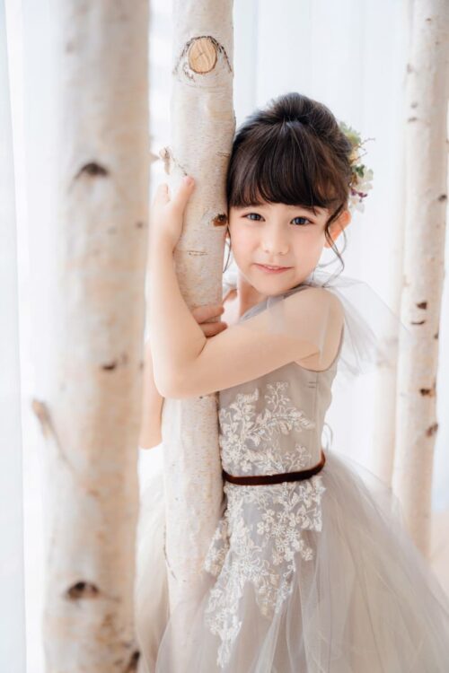 七五三の記念撮影で、グレー系のレースドレスを着た女の子が、白樺の木に寄り添いながら微笑んでいる写真