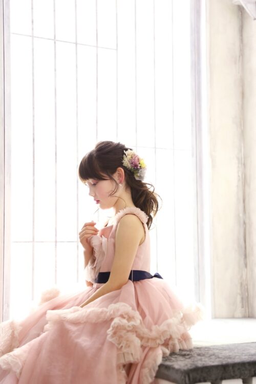 フリルと濃紺のウェストリボンが映えるピンクのドレスを着て、窓辺に腰掛け横顔を見せる七五三の女の子の写真