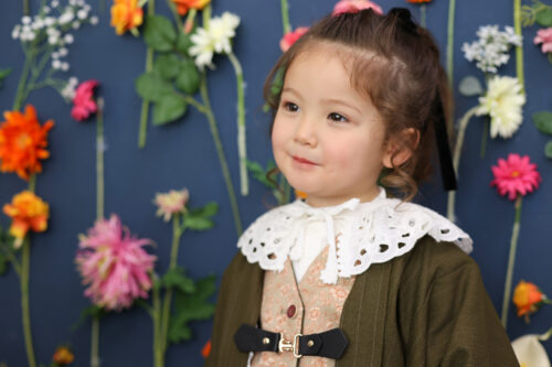 七五三の女の子の記念写真。カーキ色のクラシカルなドレスにレース襟を合わせ、色とりどりの花を背景に微笑んでいる