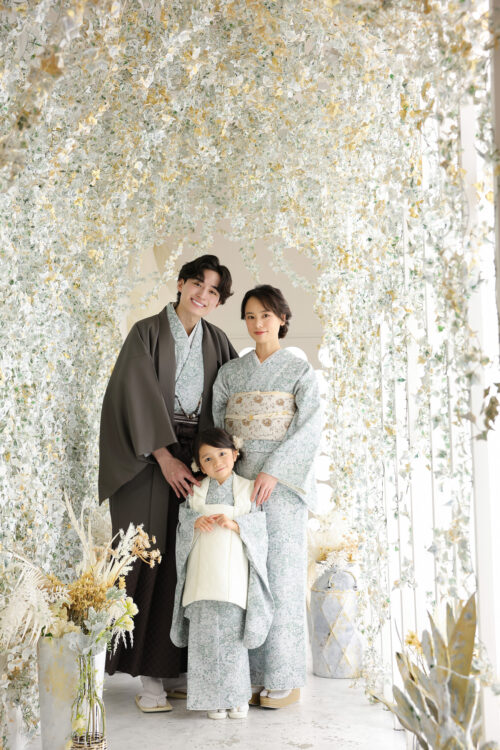七五三3歳の家族写真。着物姿の両親と娘が並んで立ち、緑のアーチが続く通路で微笑んでいる