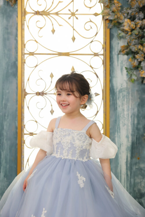 七五三3歳の女の子の記念写真。水色のプリンセスドレスを着て、金の装飾扉を背景に笑顔を見せている