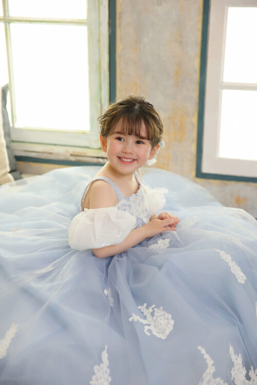 七五三3歳の女の子の記念写真。水色のプリンセスドレスを着てベッドに座り、窓辺で満面の笑顔を見せている