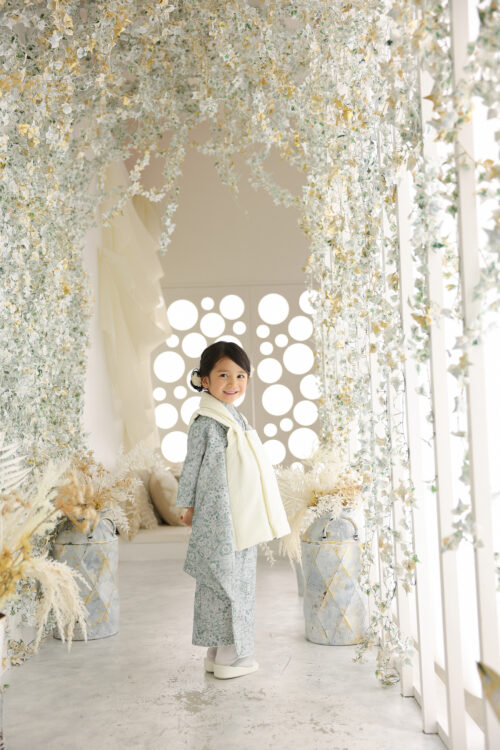 七五三3歳の女の子の記念写真。淡いグリーンの着物姿で緑のアーチが並ぶ通路に立ち、振り返って笑顔を見せている