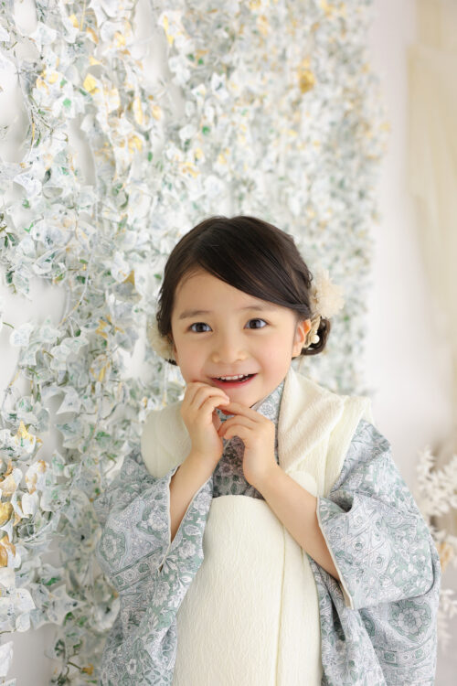 七五三3歳の女の子の記念写真。淡いグリーンの着物姿で両手をあごの下で組み、はにかんだ笑顔を見せている