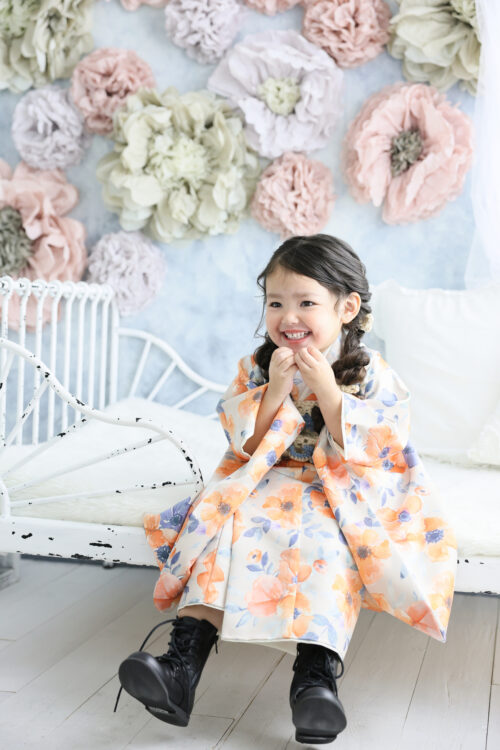 七五三3歳の女の子の記念写真。オレンジと青の花柄の着物にブーツを合わせ、ベンチに座って満面の笑顔を見せている