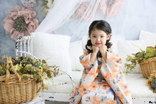 七五三3歳の女の子の記念写真。オレンジと青の花柄の着物を着て、両手にあごをのせてこちらを見つめている