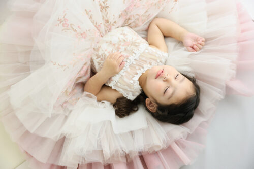 七五三3歳の女の子の記念写真。花柄が施されたピンクのチュールドレスに包まれて仰向けに寝転び、目を閉じて微笑んでいる
