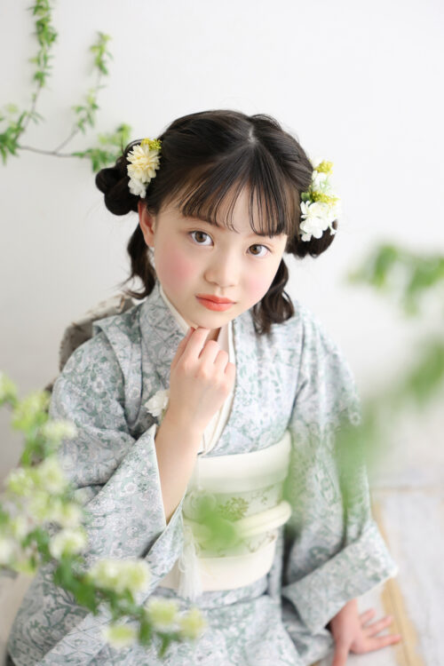 七五三7歳の女の子の記念写真。淡いグリーンの着物姿で両側に白い花飾りをつけ、あごに手を添えている