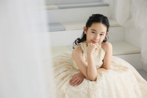 七五三7歳の女の子の記念写真。アイボリーのレースドレスを着て、あごに手を添えて優しく微笑んでいる