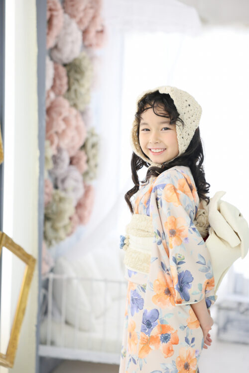 七五三7歳の女の子の記念写真。オレンジと青の花柄の着物に編み込みの帽子を合わせ、振り返って笑顔を見せている