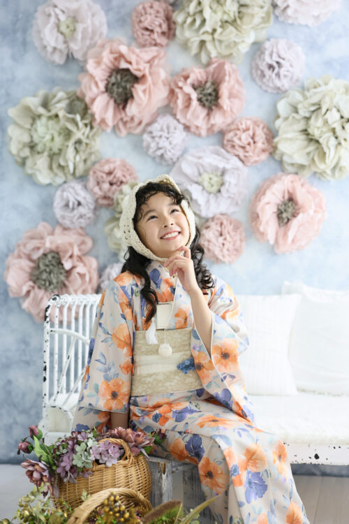 七五三7歳の女の子の記念写真。オレンジと青の花柄の着物に編み込みの帽子を合わせ、あごに手を添えて上を見上げている