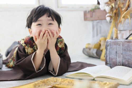 七五三の男の子の記念写真。花柄襟の羽織姿で開いた本の前にうつ伏せになり、両手で頬を包みながらはにかんだ笑顔を見せている