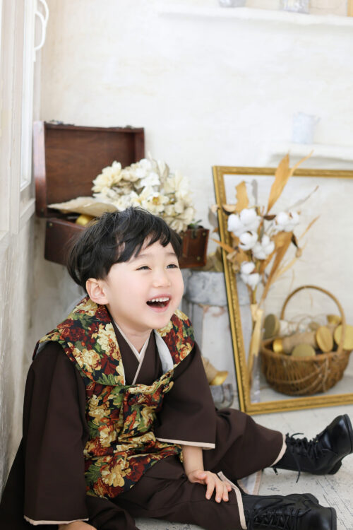 七五三の男の子の記念写真。花柄襟の羽織姿で床に座り、上を向いて大きな口を開けて笑っている