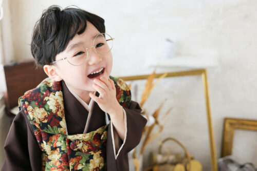 七五三の男の子の記念写真。丸眼鏡と花柄襟の羽織を合わせ、口元に手をあてて満面の笑顔を見せている