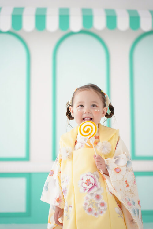 STUDIO ARCららテラスHARUMI FLAG店で撮影した七五三の女の子の記念写真。黄色の花柄着物姿で大きな飴を手に、満面の笑顔を見せている