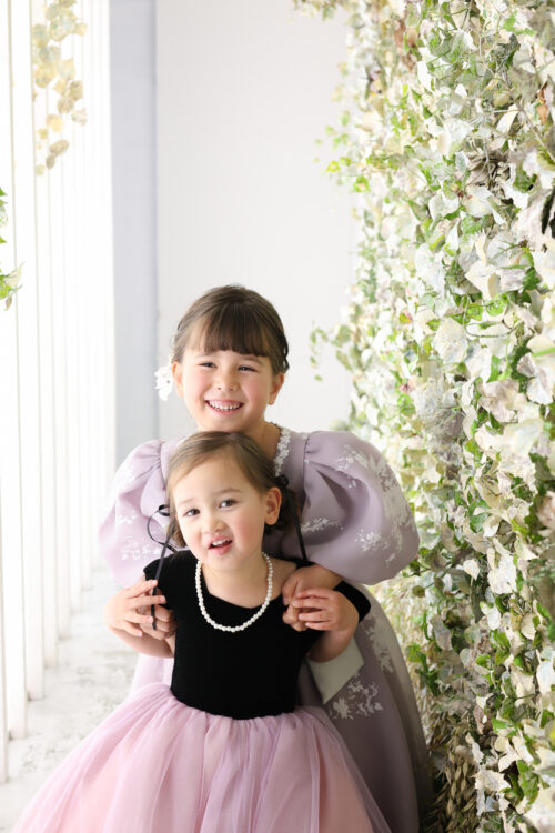 七五三3歳と5歳の姉妹の記念写真。パープルのパフスリーブドレスとピンクのチュールスカートで並び、満面の笑顔を見せている