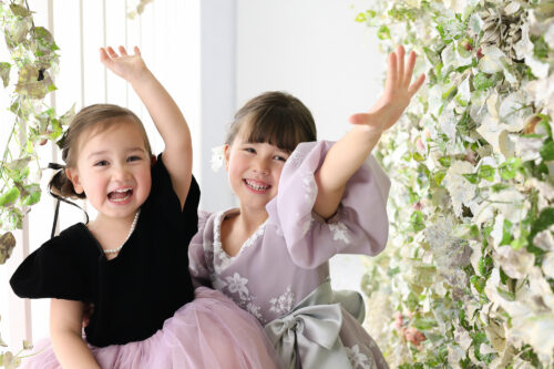 七五三3歳と5歳の姉妹の記念写真。黒とラベンダー色のドレスを着て両手を高く上げ、はじけるような笑顔を見せている