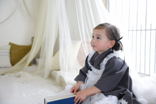 七五三3歳の女の子の記念写真。グレーのコートに白いフリル襟を合わせ、本を手に持ちながら窓の外を見つめている