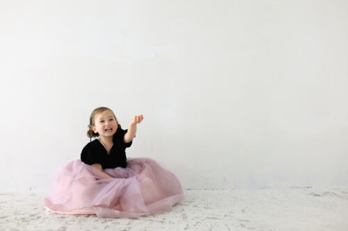 七五三3歳の女の子の記念写真。黒トップスにピンクのチュールスカートを着て、手を伸ばしながら笑顔を見せている
