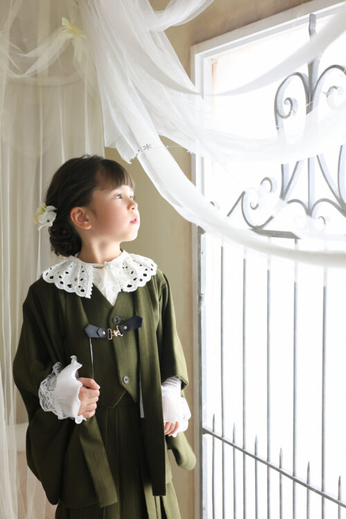 STUDIO ARC心斎橋店で撮影した七五三5歳の女の子の記念写真。カーキ色のベスト袴に白いレース襟を合わせ、窓辺で上を見上げている