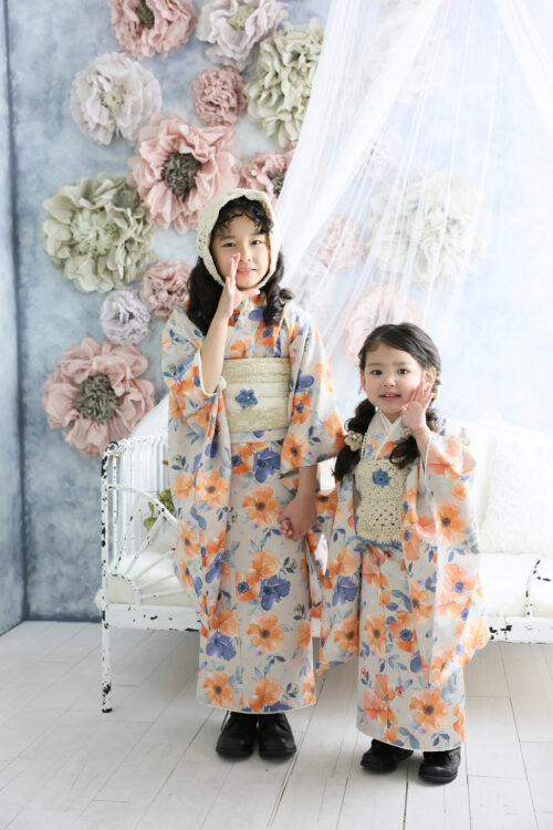 七五三3歳と7歳の姉妹の記念写真。オレンジと青の花柄のおそろいの着物姿で手をつなぎ、花の壁を背景に立っている