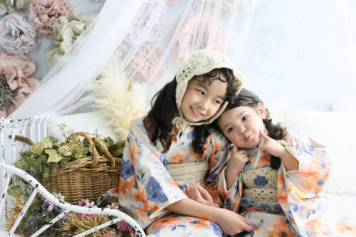 七五三3歳と7歳の姉妹の記念写真。オレンジと青の花柄のおそろいの着物を着て寄り添い、妹がおどけた表情を見せている