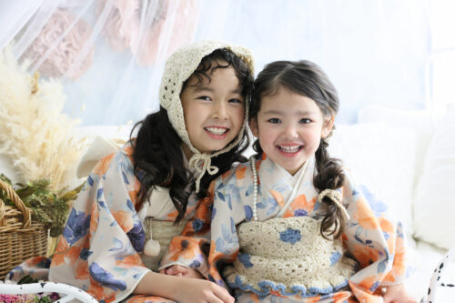 七五三3歳と7歳の姉妹の記念写真。オレンジと青の花柄のおそろいの着物を着て、満面の笑顔を見せている