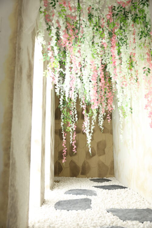 STUDIO ARC枚方店の撮影セット。天井一面から白とピンクの藤のような花飾りと緑の葉が垂れ下がり、足元には白い玉砂利の上にグレーの飛び石が奥へと続いている。突き当たりは石積み風の壁、両側はアンティーク調の白い壁で、花のトンネルをくぐるような和の雰囲気の空間。
